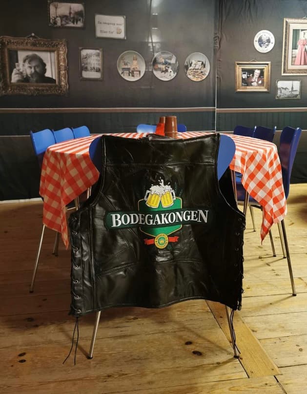 Bodegakongens lædervest med logo hænger på en stol ved et bord med ternet dug